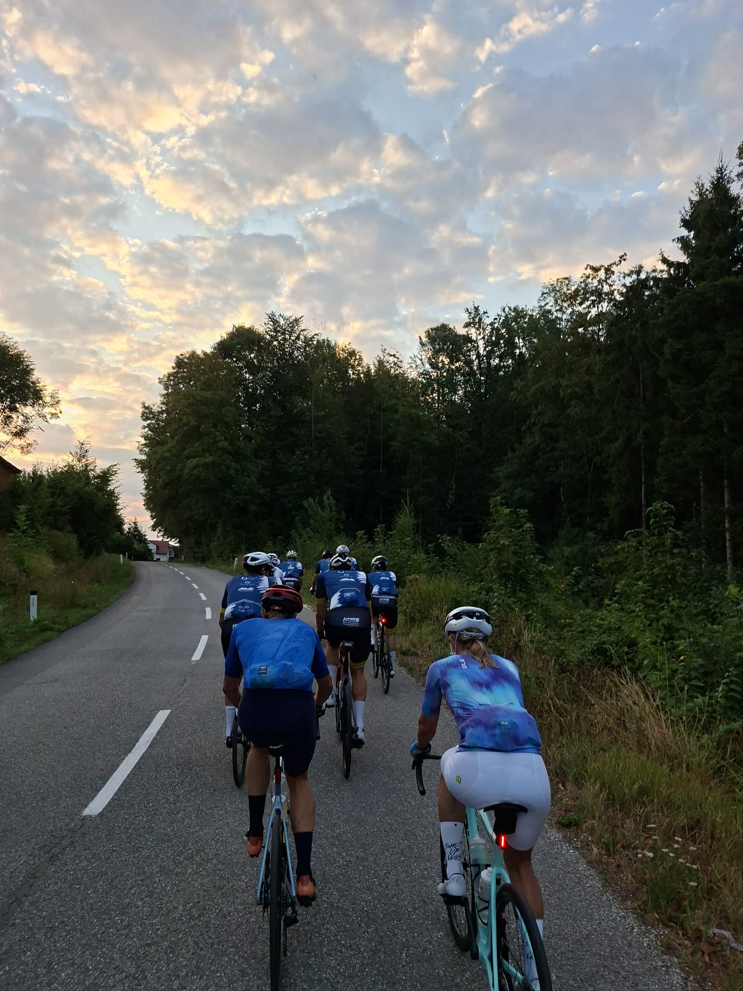 Radfahrerinnen und Radfahrer fahren auf einer asphaltierten Straße durch eine ländliche Gegend bei Sonnenuntergang.